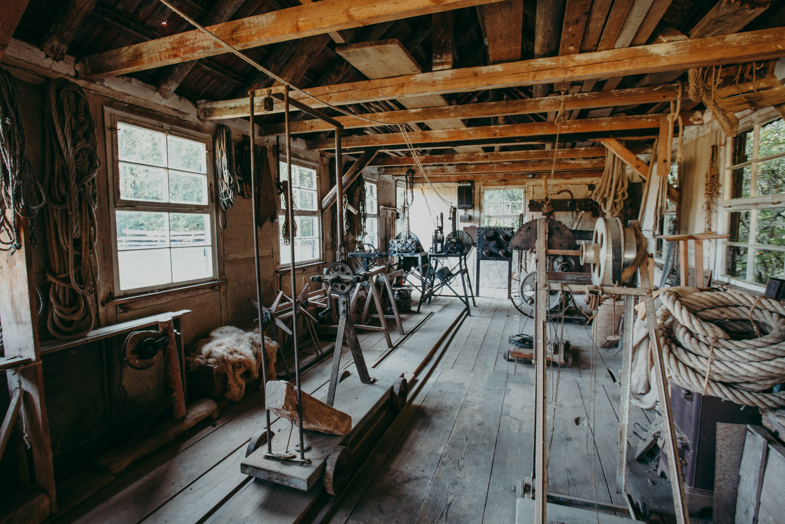 Foto: Innenraum einer historischen Seilerei mit Holzbalken, Maschinen zur Seilherstellung und mehreren Seilen, die an den Wänden hängen. Auf der rechten Seite sind große Seilrollen, Licht fällt durch die Fenster.