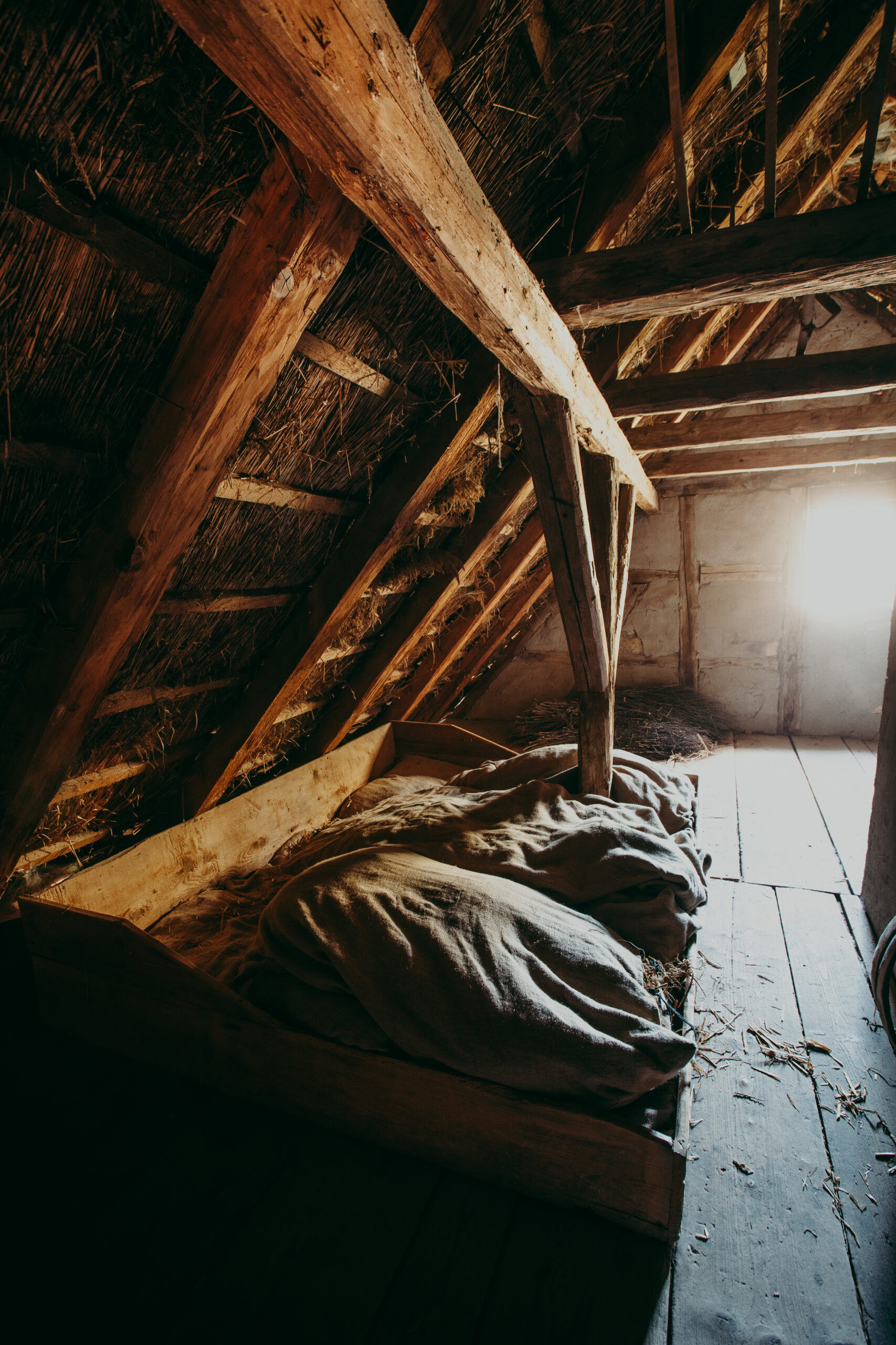 Foto: Ein niedriges, hölzernes Bettgestell mit Strohsack unter einem Reetdach. Der Raum ist nur schwach beleuchtet.