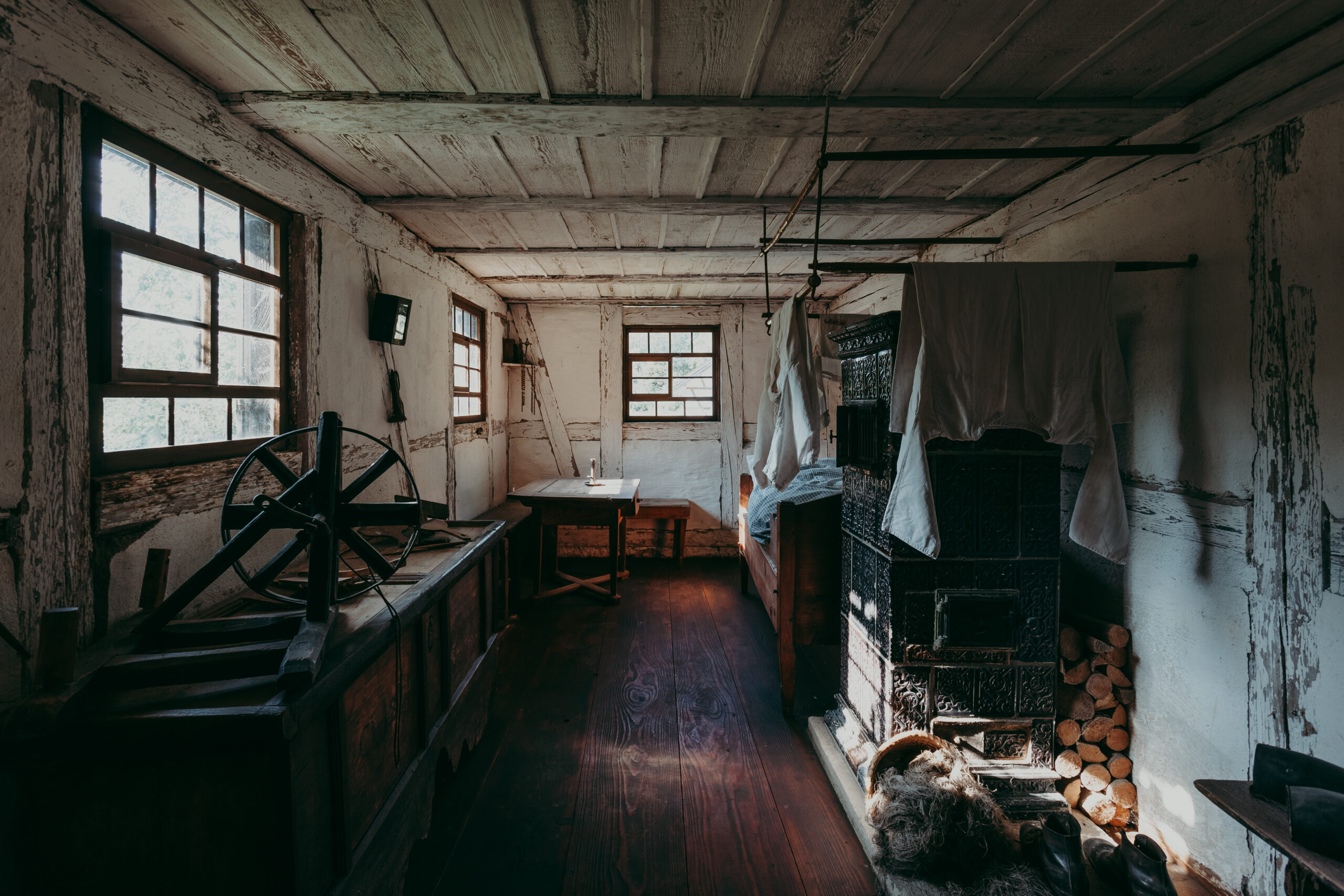 Foto: Historischer Wohnraum mit dunklem Holzfußboden. Links ein Webstuhl, rechts ein großer grüner Kachelofen, darüber trocknende Wäsche. Dahinter steht in der Ecke ein Bett. In der Mitte ein Tisch mit Stuhl. Tageslicht fällt durch mehrere kleine Fenster.