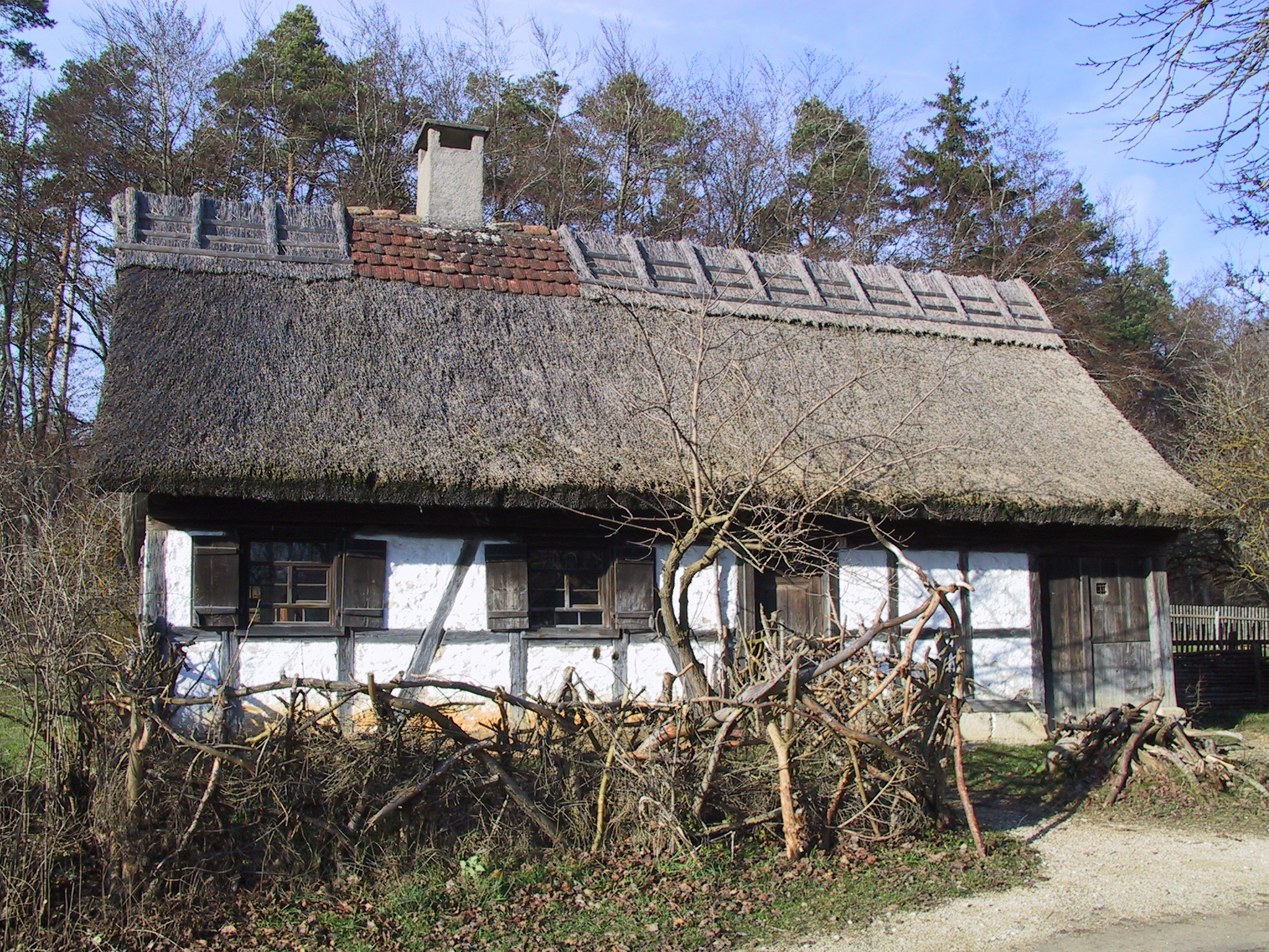 Foto: Kleines Fachwerkhaus mit Reetdach, weißen Gefachen und dunklem Holz. Davor ein niedriger Zaun aus geschnittenem Geäst.