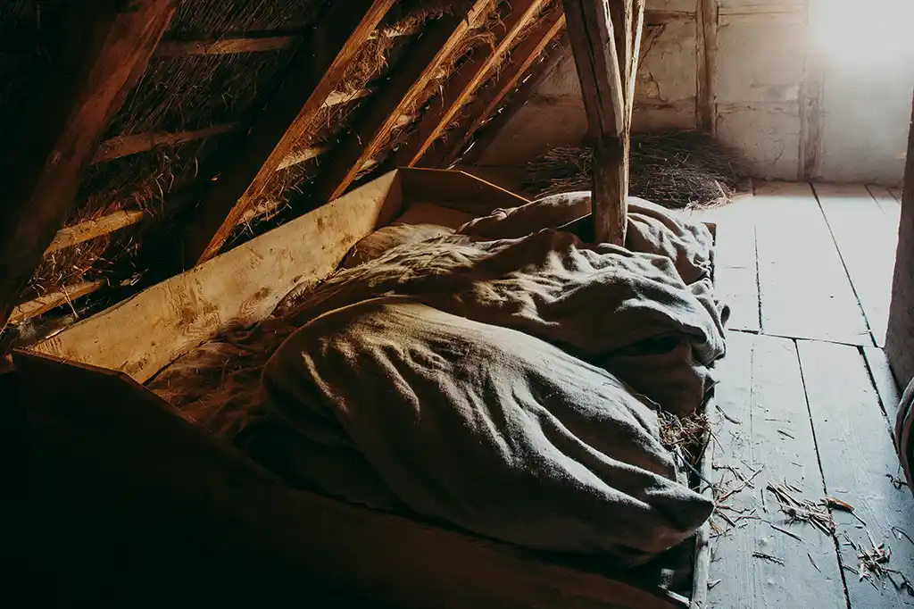 Foto: Ein niedriges, hölzernes Bettgestell mit Strohsack unter einem Reetdach. Der Raum ist nur schwach beleuchtet.