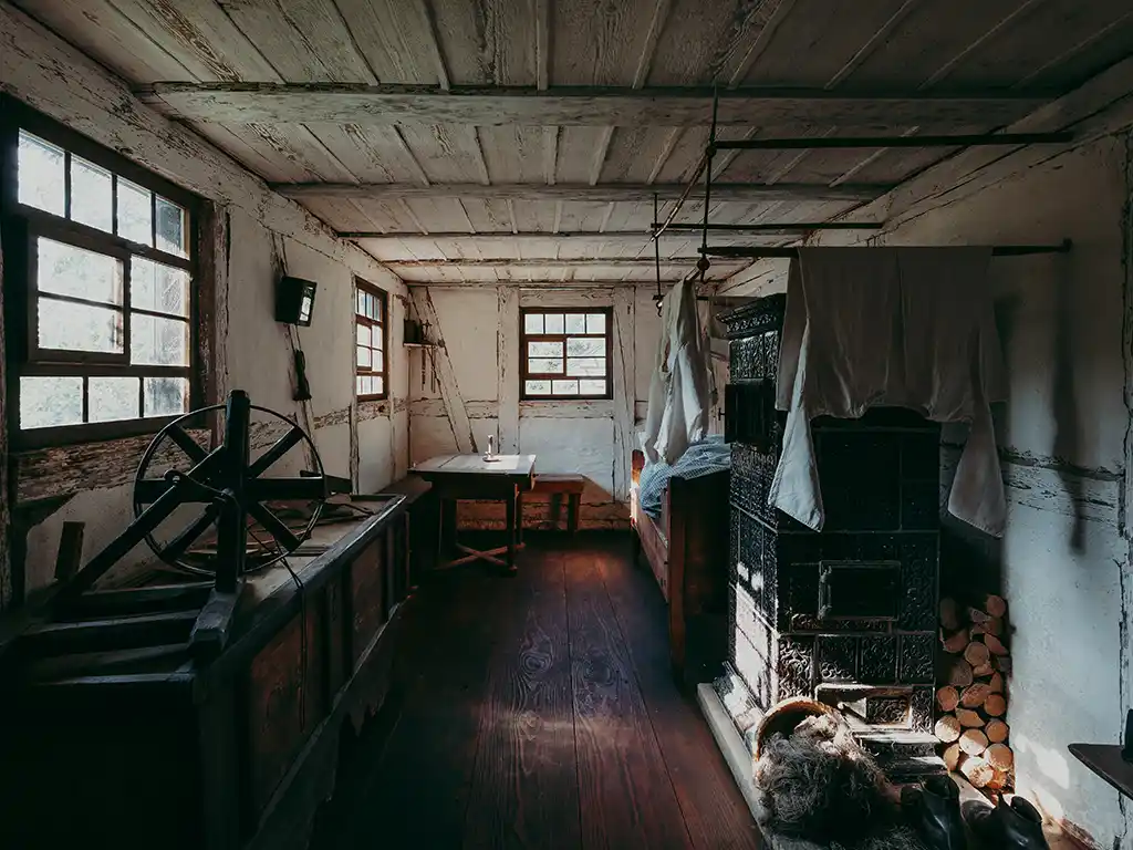 Foto: Historischer Wohnraum mit dunklem Holzfußboden. Links ein Webstuhl, rechts ein großer grüner Kachelofen, darüber trocknende Wäsche. Dahinter steht in der Ecke ein Bett. In der Mitte ein Tisch mit Stuhl. Tageslicht fällt durch mehrere kleine Fenster.