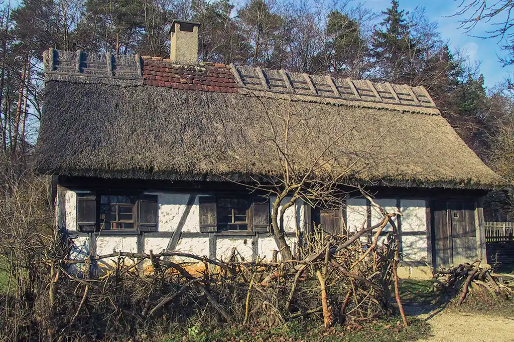 Foto: Kleines Fachwerkhaus mit Reetdach, weißen Gefachen und dunklem Holz. Davor ein niedriger Zaun aus geschnittenem Geäst.