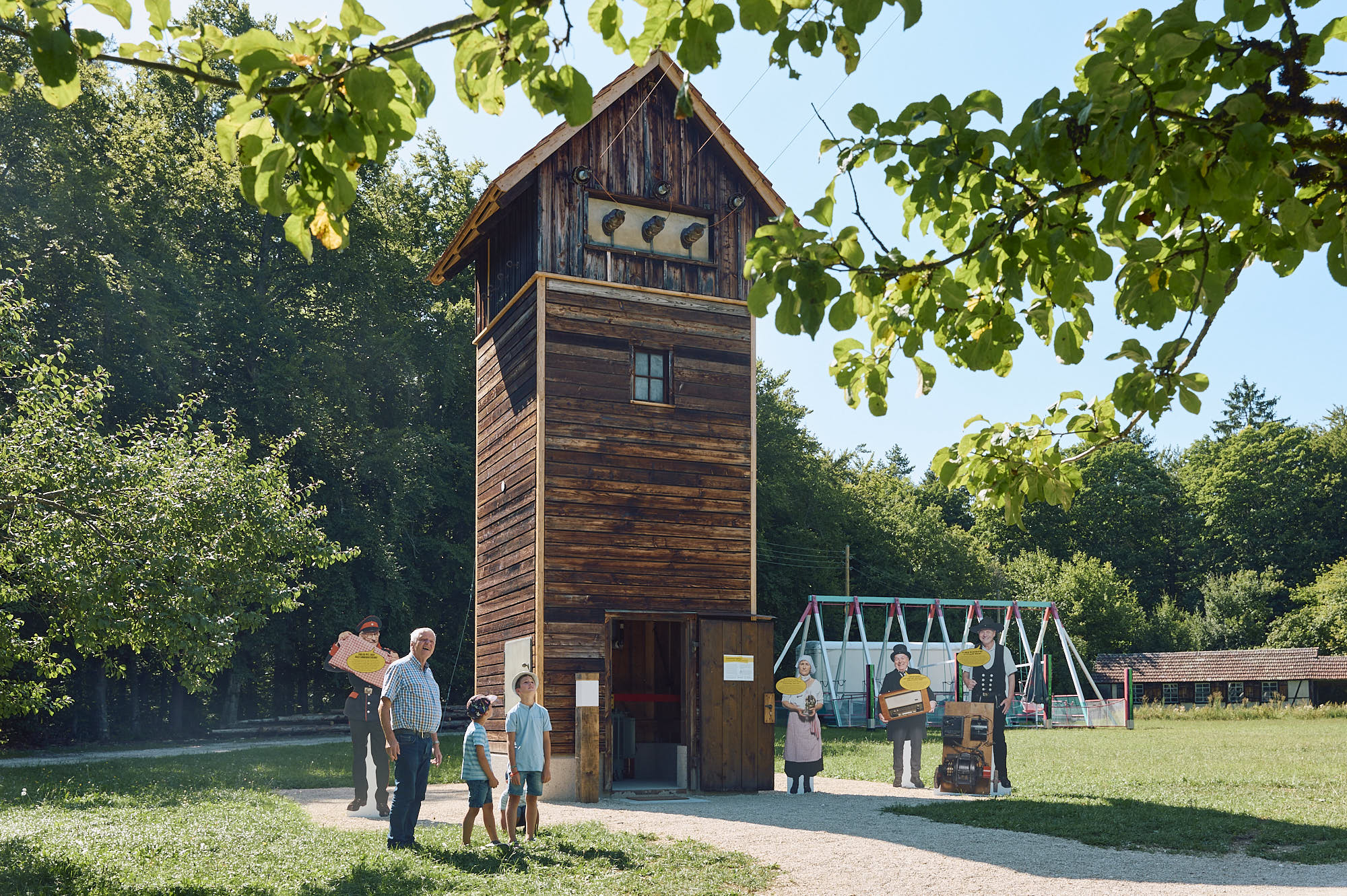 Foto: Drei Kinder und ein älterer Mann stehen vor. einer holzverkleideten Umspannstation, dessen Türen geöffnet sind. Daneben sind Aufsteller von Personen in historischer Kleidung.