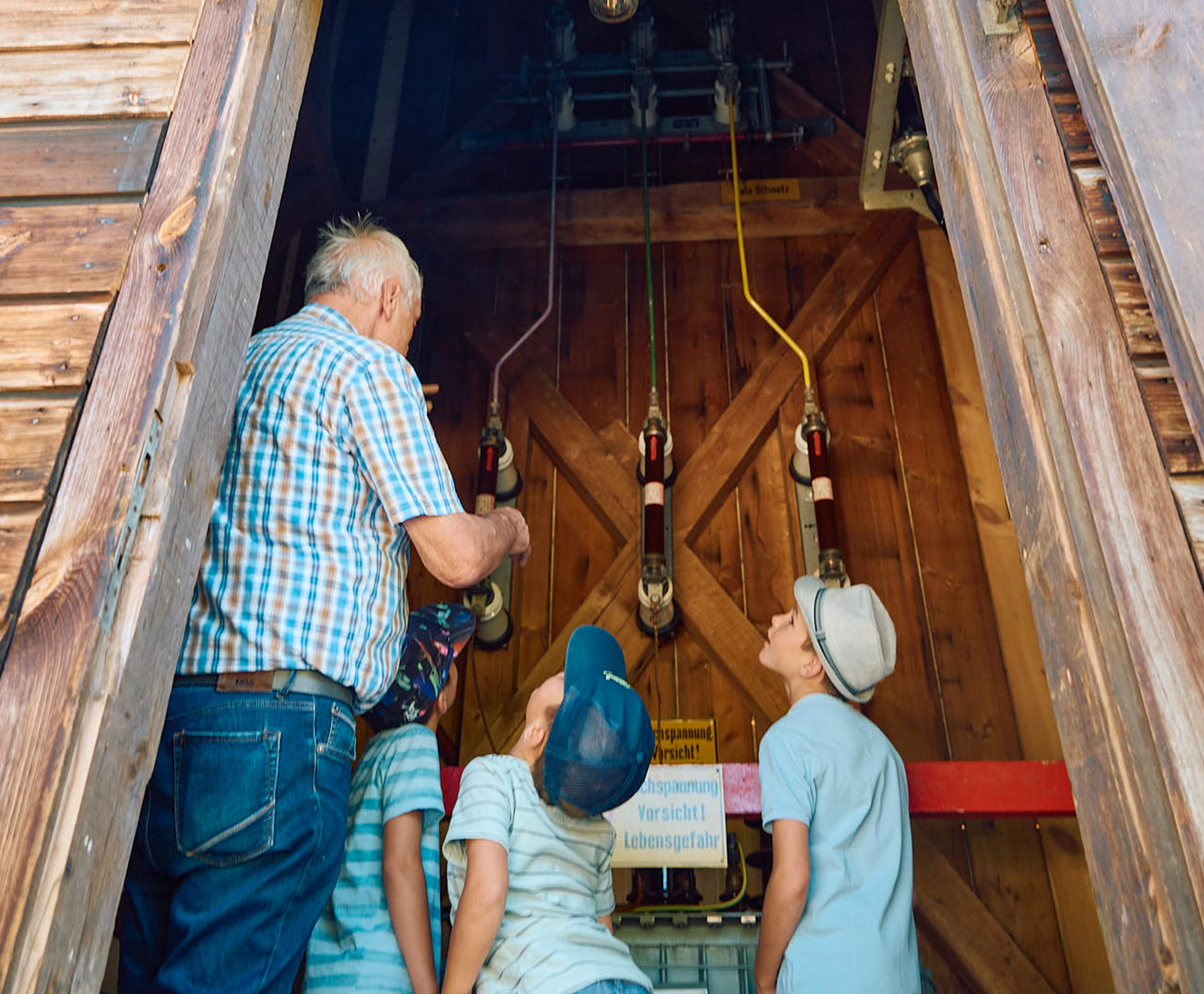 Foto: Blick in den Innenraum der Umspannstation: Ein älterer Mann und drei Kinder betrachten gemeinsam die elektrischen Bauteile an der Wand des hölzernen Turminneren. Warnschild mit der Aufschrift: Hochspannung – Vorsicht Lebensgefahr.