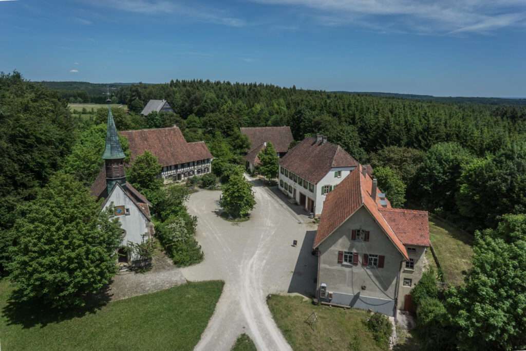 Luftaufnahme des Freilichtmuseums Neuhausen ob Eck mit Blick auf den Dorfplatz. Mehrere historische Gebäude mit roten Ziegeldächern gruppieren sich um einen offenen Platz, umgeben von Bäumen und Waldlandschaft.